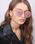 A woman with long brown hair models For Art's Sake® T-shirt - Sunglasses, featuring pink-tinted UVA & UVB protection, along with gold hoop earrings and a black leather jacket against a light gray background.