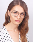 A woman with brown hair and a neutral expression is wearing geometric eyeglasses with heptagon-shaped lenses called Antidote by For Art's Sake® and large hoop earrings. She is dressed in a white blouse with black polka dots, standing against a plain background.