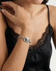 A person in a black lace-trimmed camisole showcases The Geometry Bracelet by For Art's Sake®, featuring a large square centerpiece. Their other hand touches their shoulder, enhancing the bracelet's modern, stylish appeal.