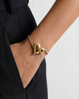 A person in black pants with a hand in their pocket showcases For Art's Sake® The Puffy Heart Bracelet, featuring a gold chain and two puffed heart charms, against a plain light background.
