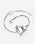 The Puffy Heart Bracelet by For Art's Sake® is a silver chain bracelet with a shiny, abstract puffed heart pendant and an adjustable clasp featuring a circular FAS charm, shown on a white background.