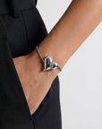 A person in black pants slips a hand into their pocket, displaying For Art's Sake® The Puffy Heart Bracelet—a delicate silver chain with two polished puffed heart charms and a subtle FAS charm on their wrist.