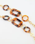 A close-up view of a stylish chain necklace featuring alternating large tortoiseshell oval links and smaller circular links, connected by 18-karat gold-plated rings. The NYC Glasses Chain by For Art's Sake® has a sophisticated, modern design with oversized acetate links and is displayed against a plain white background.