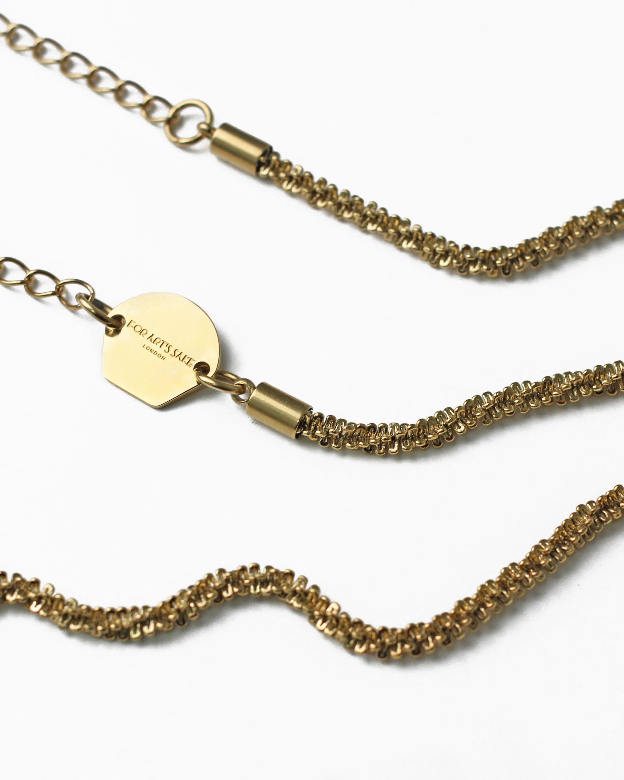 Close-up of an 18k gold-plated Mayfair Glasses Chain by For Art's Sake® featuring intricate woven links. The chain has an adjustable, heart-shaped claw closure and a small hexagonal tag engraved with the words "FOR ART'S SAKE." The background is white, highlighting the exquisite details of the chain and tag.