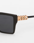 Close-up of Castle by For Art's Sake®: black oversized square frames with a gold chain-link detail on the arm, crafted from lightweight acetate, set against a plain white background.