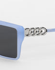 Close-up of For Art's Sake® Castle sunglasses with blue oversized square acetate frames, dark lenses, and a silver chain-link detail on the temple, set against a plain white background.