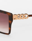 Close-up of For Art's Sake® Castle oversized square frames in tortoiseshell, made from lightweight acetate with gold chain details on the arms and brown gradient lenses, set against a white background.