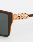 Close-up of Castle sunglasses by For Art's Sake® featuring oversized square frames in brown and gold lightweight acetate with chain details on the temples, set against a plain white background.