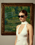 A woman in a white halter dress and For Art's Sake® Claude Monet (Limited Edition) sunglasses stands before a framed painting of a garden bridge over a pond, evoking Monet's Water Lilies.