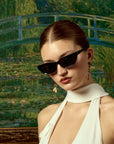 A woman with slicked-back hair models For Art's Sake® Claude Monet (Limited Edition) black cat-eye sunglasses and gold earrings, wearing a white halter top before a dreamy garden scene with a bridge and water lilies.