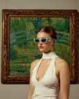A woman in a chic white dress wears For Art's Sake® Claude Monet (Limited Edition) sunglasses, standing before a framed impressionist painting of a lush garden and arched bridge inspired by Monet.