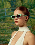 A woman in a white halter dress and sleek bun wears For Art's Sake® Claude Monet (Limited Edition) cat-eye sunglasses and dangling earrings, standing before a painting of a bridge over a lily pond.