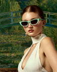 A woman in a cream halter dress and neat updo poses by Monet’s Japanese bridge, wearing For Art's Sake® Claude Monet (Limited Edition) sunglasses and bold gold earrings.