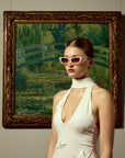 A woman wearing For Art’s Sake® Claude Monet (Limited Edition) sunglasses and a white dress stands before a framed Monet Japanese Bridge, blending modern style with timeless art.