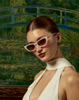 A stylish woman in For Art's Sake® Claude Monet (Limited Edition) cat-eye sunglasses and a white halter dress poses before Monet’s Japanese Bridge, her hair slicked back and gold earrings glinting.