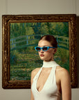 A woman in a white sleeveless dress wears For Art's Sake® Claude Monet (Limited Edition) sunglasses, posing before a framed Monet-style garden bridge painting—fusing modern fashion with classic art.