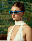 A woman in For Art’s Sake® Claude Monet (Limited Edition) cat-eye sunglasses and a white halter dress stands before a green garden bridge painting, her hair pulled back and wearing long, dangling earrings.