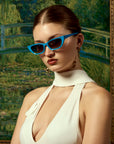 A woman poses in front of a Monet-inspired garden bridge painting, wearing For Art's Sake® Claude Monet (Limited Edition) cat-eye sunglasses and a white halter dress, her hair up with gold earrings.