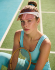 A young woman in a blue athletic outfit wears the For Art's Sake® Cloud Visor with an adjustable elastic strap and UV protection, sitting on a tennis court with a wooden racket. Green court lines appear in the background.
