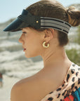 A woman wears the For Art's Sake® Cloud Visor (UV protected, black) and gold hoop earrings with her hair in a low bun. She dons a colorful leopard print garment and stands outdoors, looking off to the side in bright sunlight.