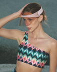 A woman outdoors wears a UV-protected Cloud Visor by For Art's Sake® and a patterned one-shoulder crop top, shading her eyes as she looks into the distance.