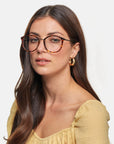 A woman with long brown hair is wearing For Art's Sake® Club tortoiseshell glasses that feature a blue light filter and gold hoop earrings. She has a calm expression and is dressed in a light yellow, gathered-top blouse. The background is plain white.