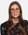 A woman with long, wavy brown hair is wearing Club glasses from For Art's Sake®, which are large and round with black frames that feature a blue light filter. She has a neutral expression and is dressed in a black sequin top. The background is plain light grey.