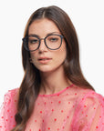 A woman with long brown hair is wearing the Club glasses from For Art's Sake®, featuring large black frames with a blue light filter, and a pink sheer top adorned with red polka dots. She has a neutral expression and is looking slightly to the side against a white background.