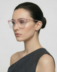 A woman with fair skin and dark hair in a low bun models the Dion pink cat-eye glasses by For Art's Sake®, paired with a one-shoulder dark gray top, against a plain light background.