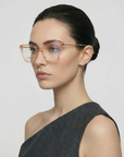 A woman with fair skin and dark hair in a low bun wears For Art's Sake® Dion oversized translucent pink cat-eye glasses with UV protection and a sleeveless asymmetrical gray top, posing against a plain light background.
