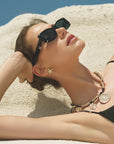 A woman reclines on a sandy rock under a clear blue sky, wearing For Art's Sake® Dyer luxury black sunglasses, starfish earrings, a gold necklace, and a black swimsuit.