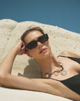 A woman with short hair reclines on a light rock under a clear blue sky, looking relaxed and stylish as she wears For Art's Sake® Dyer luxury black sunglasses and a gold medallion necklace with a black top.