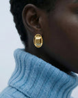 A close-up shows a person wearing The Geometry Earrings by For Art's Sake®—modern, chunky gold earrings—paired with a light blue ribbed turtleneck against a plain background.