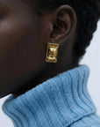 Close-up of a person wearing For Art's Sake® The Geometry Earrings—chunky, rectangular statement earrings—paired with a light blue ribbed turtleneck; only the lower face, ear, and neck are visible to highlight the modern design.