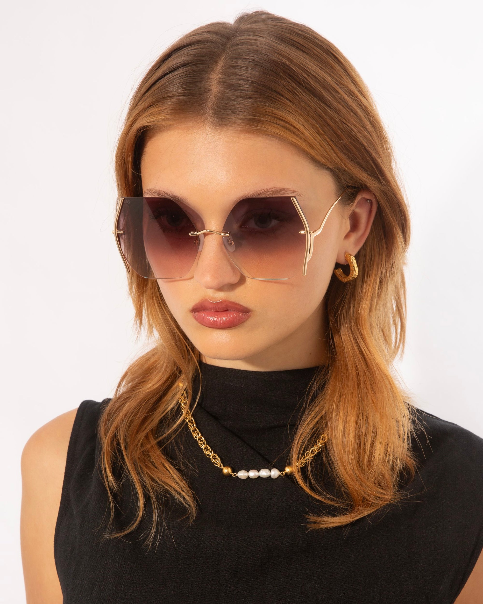 A young woman with medium-length reddish-brown hair wears large, hexagonal sunglasses called Generation by For Art's Sake® with UV protection and gradient lenses in gold-plated stainless steel frames. She has a serious expression and is dressed in a black sleeveless top, accessorized with gold hoop earrings and a gold chain necklace with a pearl.