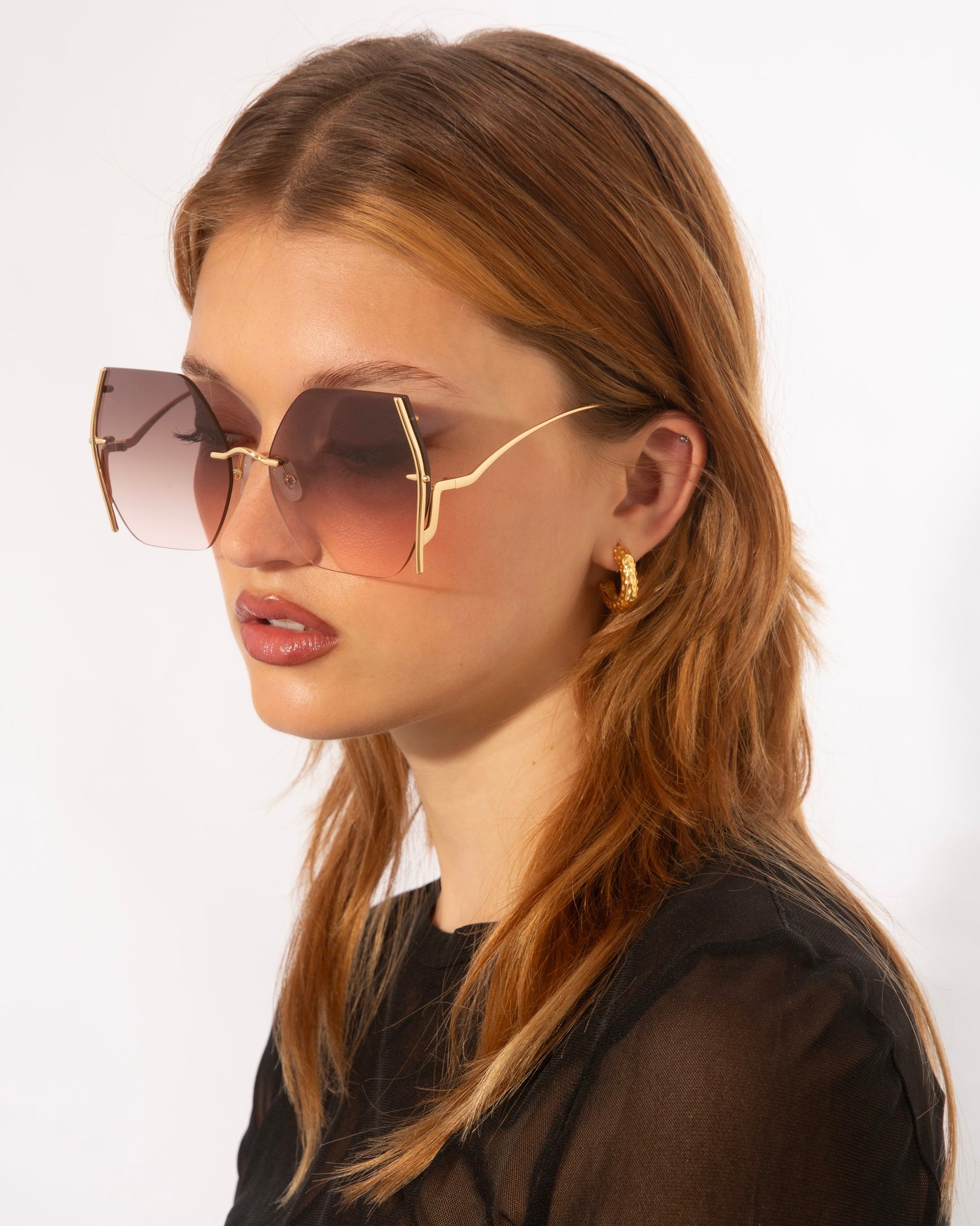 A person with long, light brown hair is wearing large, square-shaped For Art's Sake® Generation sunglasses with gold accents and gradient lenses offering UV protection. They are also wearing a black, semi-transparent top and a gold hoop earring. The background is plain white.