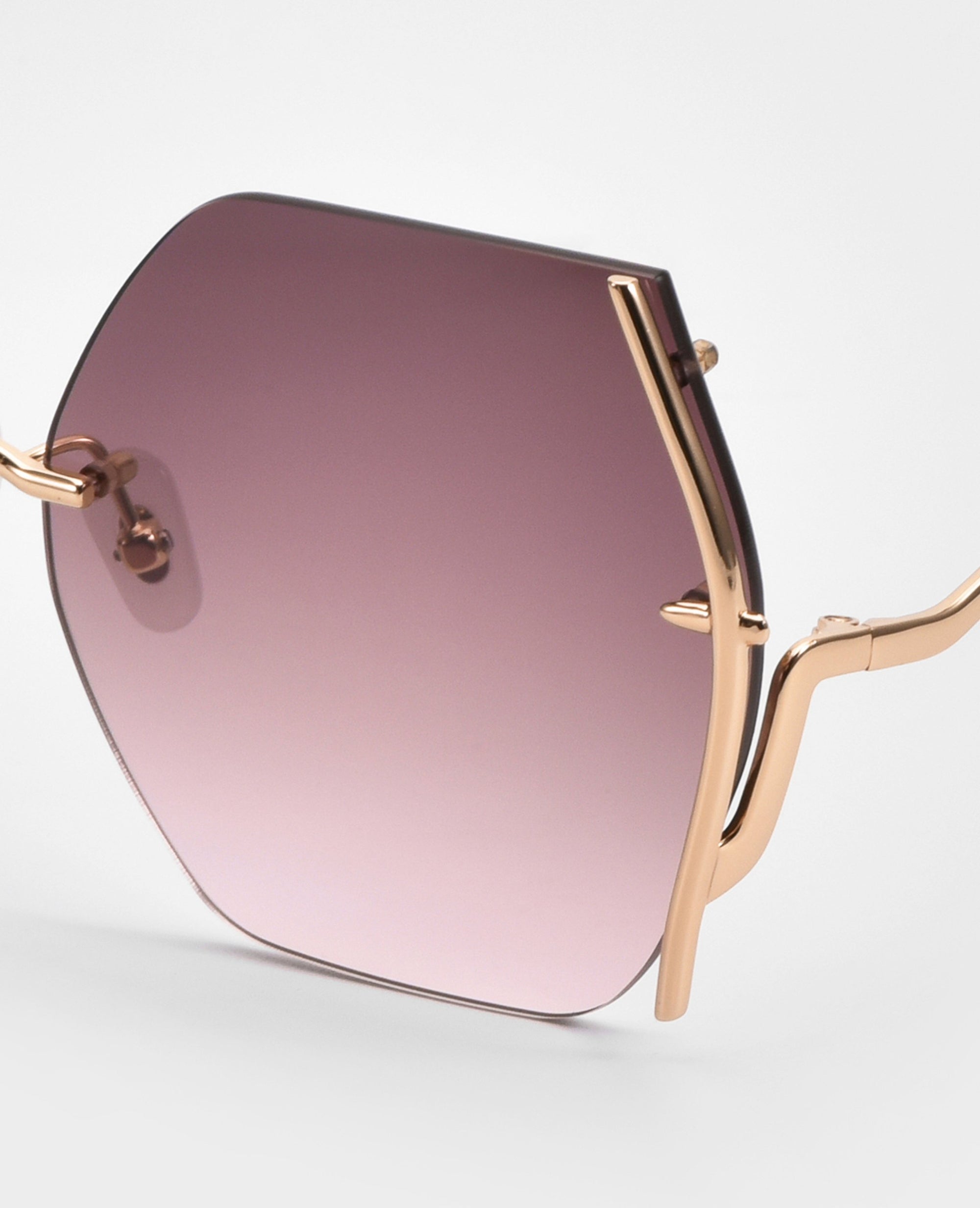 Close-up of For Art's Sake® Generation sunglasses showing a single hexagonal lens with UV protection, a pink-to-purple gradient tint, and slim gold wavy arms, set against a plain white background.