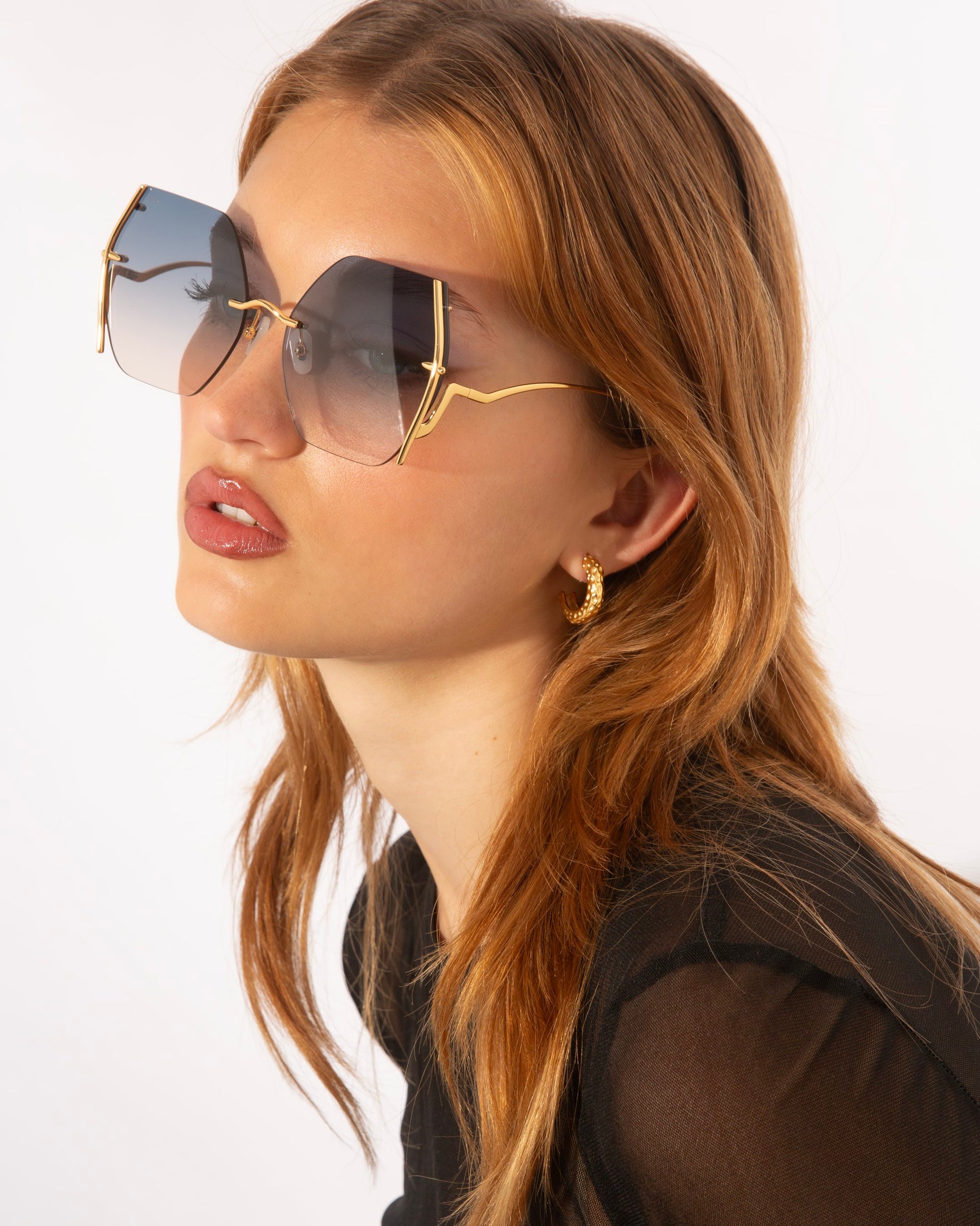 A woman with long, light brown hair wears the For Art's Sake® Generation Topaz butterfly-shaped sunglasses, featuring gold frames and UV-protective gradient lenses. She pairs them with a black sheer top and gold hoop earrings. Only 1 available.