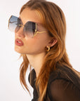 A woman with long, light brown hair wears the For Art's Sake® Generation Topaz butterfly-shaped sunglasses, featuring gold frames and UV-protective gradient lenses. She pairs them with a black sheer top and gold hoop earrings. Only 1 available.