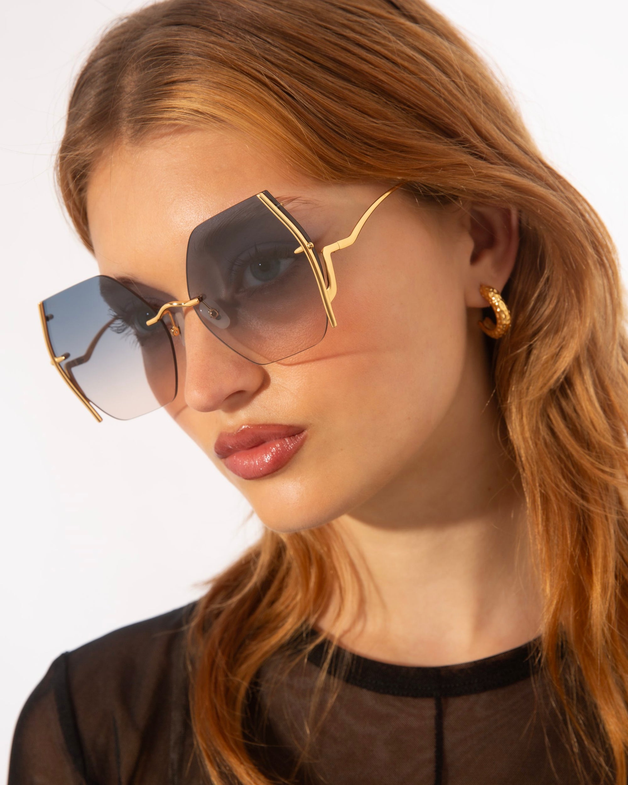 A woman with long light brown hair wears oversized For Art's Sake® Generation Topaz sunglasses (ONLY 1 AVAILABLE) with UV-protected gradient lenses and gold accents, styled with gold hoop earrings and a black sheer top for a rare find look.