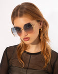 A woman with long, light brown hair wears For Art's Sake® Generation Topaz sunglasses (ONLY 1 AVAILABLE) with UV protection, gold hoop earrings, and a sheer black top. She poses against a white background—a true rare find.
