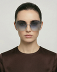 A person with straight dark hair pulled back, wearing For Art's Sake® Generation large geometric gradient sunglasses for UV protection and a dark brown top, faces forward against a plain light background.