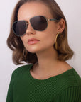 A woman with wavy light brown hair is wearing For Art's Sake® Explorer sunglasses that offer 100% UV protection, large hoop earrings with 18-karat gold plating, and a green ribbed top. She is posing against a plain background, looking slightly to the side with a neutral expression.