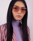 A woman with long straight black hair wears For Art's Sake® Explorer pink-tinted aviator sunglasses (worn by Beyoncé), paired with a lilac turtleneck and pink pinstripe blazer, posing confidently against a light background.