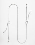 This image shows a long, delicate Florentina Glasses Chain by For Art's Sake®. The chain features two small geometric pendants and two dangling ends, each embellished with a small silver or pearl-like bead. The necklace is laid out flat against a plain white background.