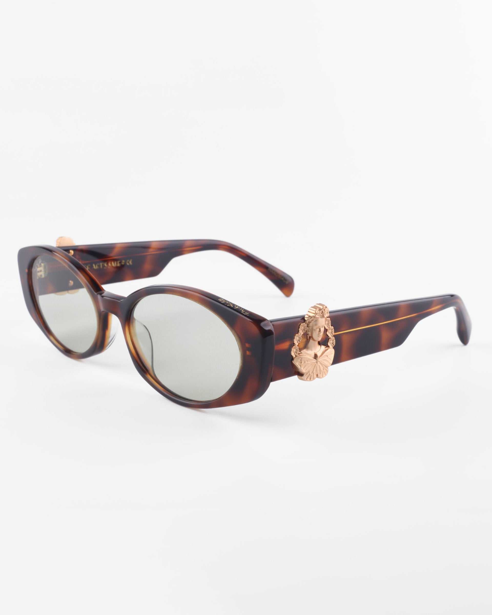 A pair of stylish Frida Portrait sunglasses by For Art's Sake® with oval lenses and tortoiseshell frames. The sunglasses feature decorative 18-karat gold-plated lion head embellishments on the temples near the hinges. The ultra-lightweight nylon lenses offer 100% UVA & UVB protection and are tinted in a light gray shade. The background is plain white.