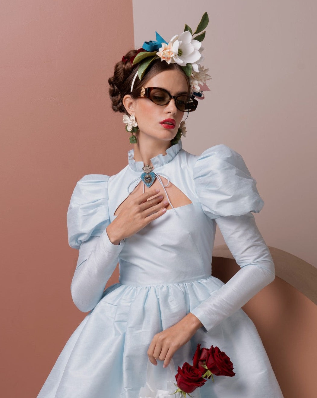 A woman dressed in a light blue vintage-style dress with puffed sleeves poses against a color-blocked background. She wears floral headgear, ultra-lightweight For Art's Sake® Frida Portrait sunglasses with 100% UVA & UVB protection, drop earrings, and holds red roses in one hand while touching her chest with the other.