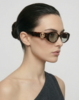 A woman with dark hair in a low bun models the For Art's Sake® Frida Portrait cateye tortoiseshell sunglasses with gold detail and a one-shoulder dark gray top, set against a plain light background.