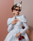 A woman dressed in a light blue vintage-style dress with puffed sleeves poses against a color-blocked background. She wears floral headgear, ultra-lightweight For Art's Sake® Frida Portrait sunglasses with 100% UVA & UVB protection, drop earrings, and holds red roses in one hand while touching her chest with the other.