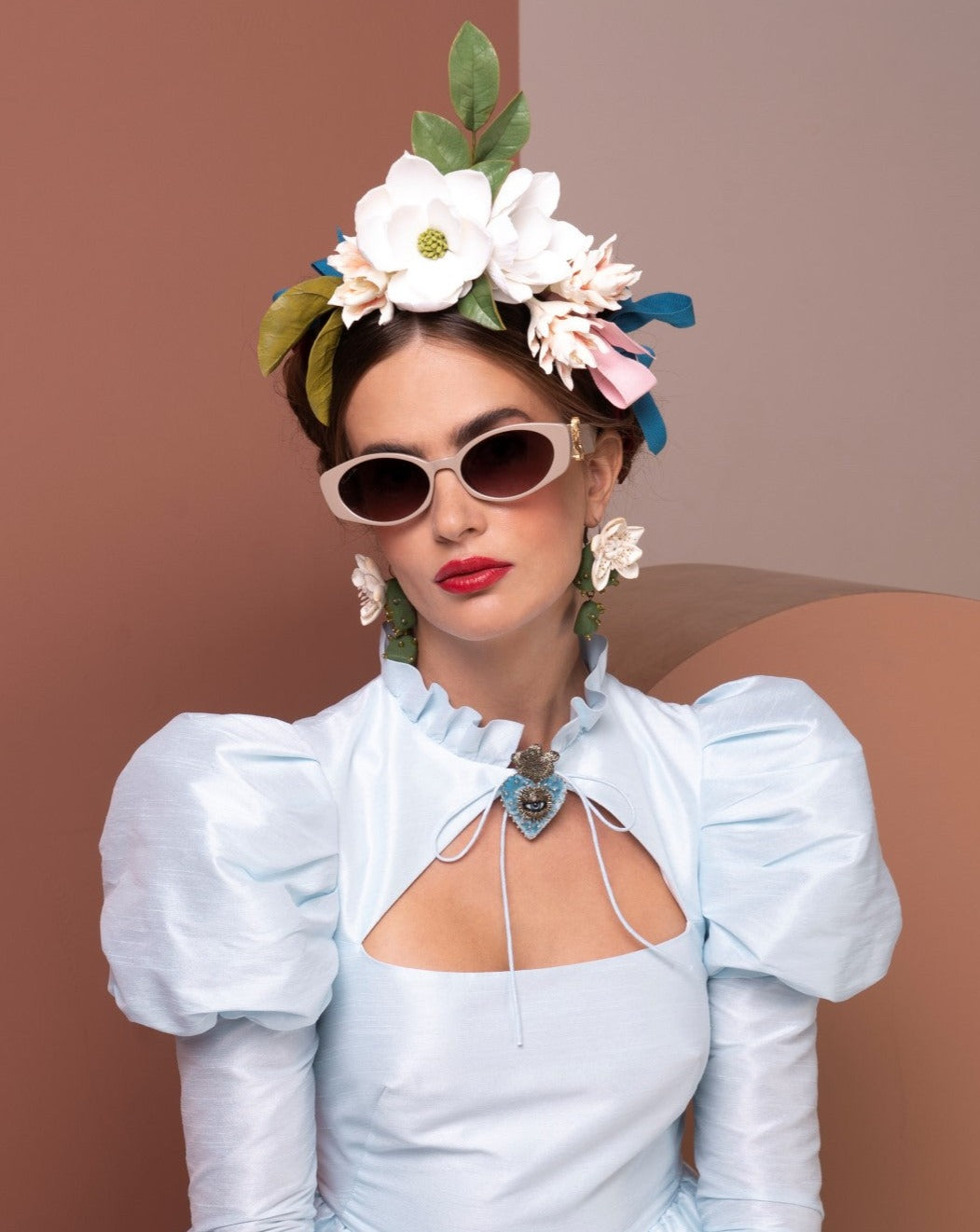 A woman wearing a light blue dress with puffed sleeves, white Frida Portrait by For Art's Sake® sunglasses with ultra-lightweight nylon lenses, and flower earrings poses confidently. She has a floral headpiece adorned with various flowers and leaves atop her dark hair. The background features earthy tones, enhancing her radiant look.